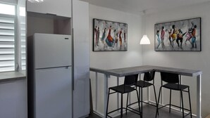 Une cuisine compacte avec un réfrigérateur blanc, un plan de travail gris et deux tabourets de bar noirs. Deux tableaux abstraits sont accrochés au mur.