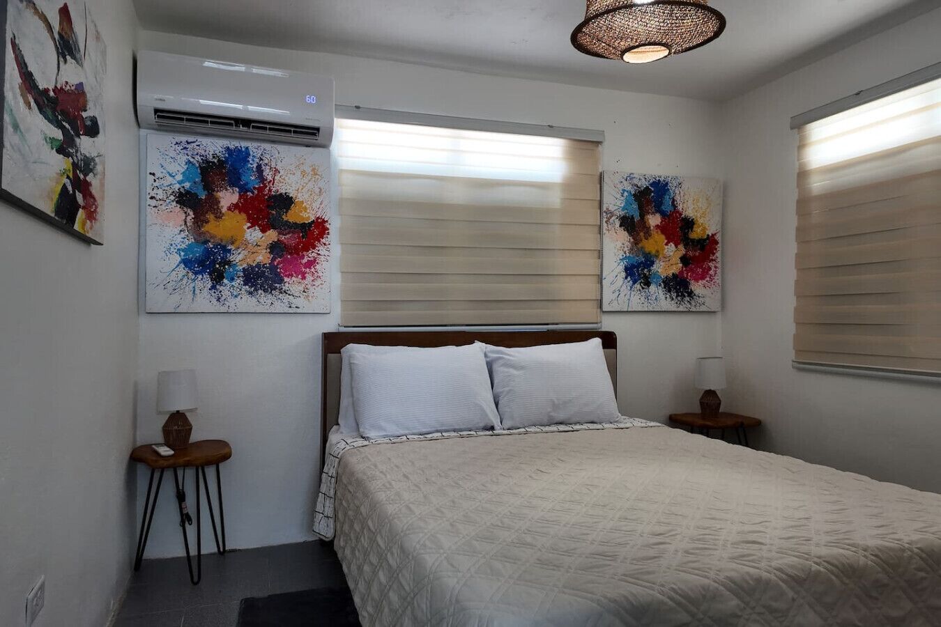 Un dormitorio con una cama, dos mesitas de noche, aire acondicionado instalado en la pared y dos cuadros abstractos.