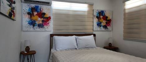 A bedroom with a bed, two bedside tables, a wall-mounted air conditioner, and two abstract paintings.