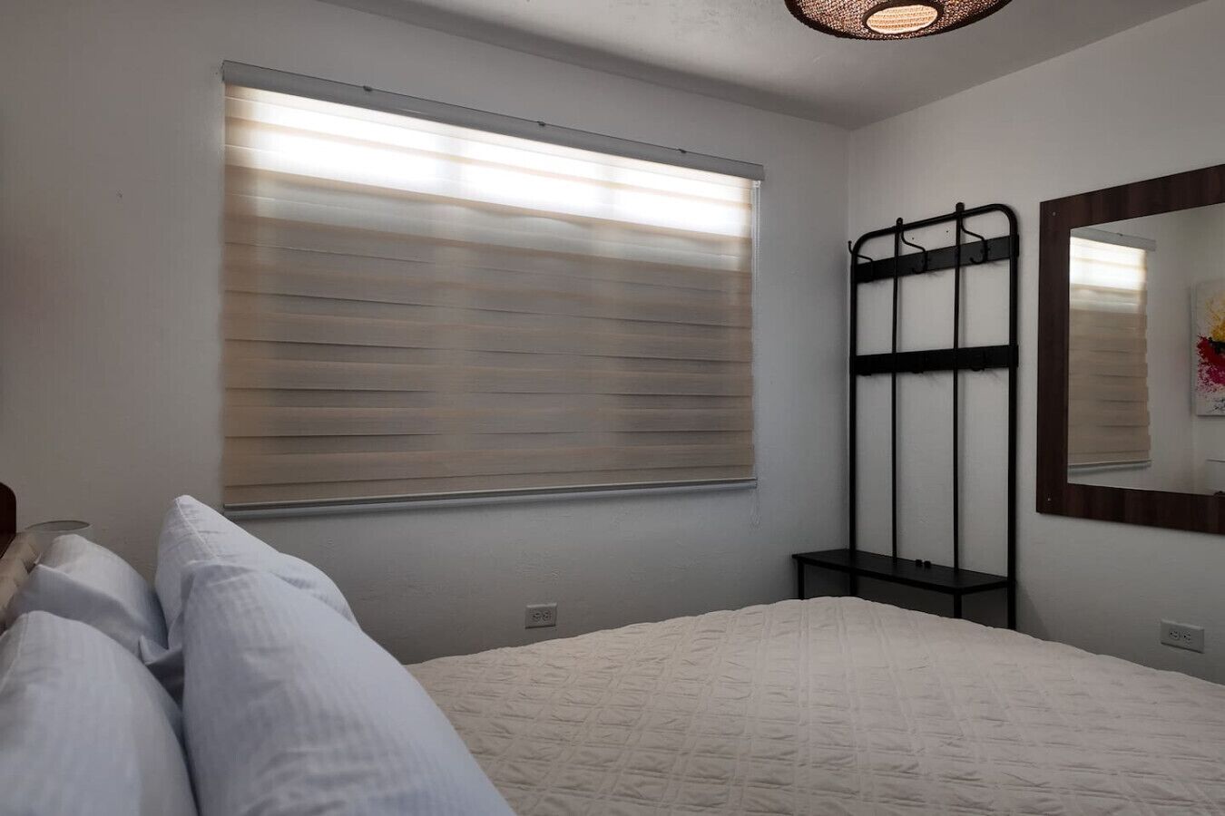 A bedroom with a bed, a window with blinds, a black metal bed frame, a bedside table, and a mirror on the wall.