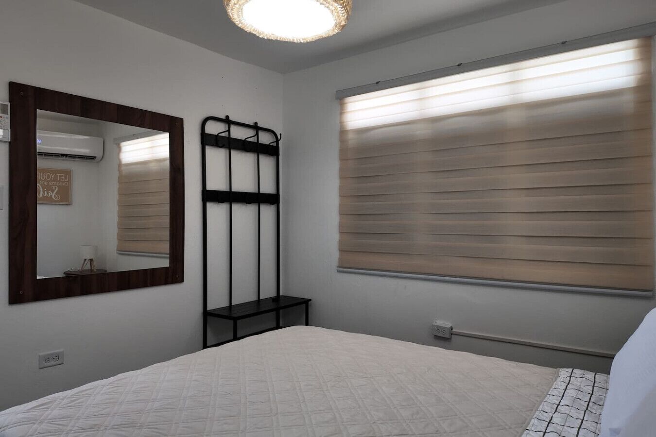 A bedroom with a bed, a mirror, an air conditioning unit, a window with blinds, and a bedside table.