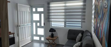 Un salon avec un canapé gris, une petite table avec une lampe, un ventilateur de plafond et une porte avec des stores.