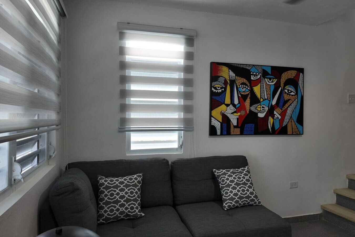 A living room with a gray sofa, patterned pillows, a window with blinds, and a colorful abstract painting on the wall.