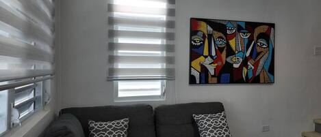 A living room with a gray sofa, patterned pillows, a window with blinds, and a colorful abstract painting on the wall.
