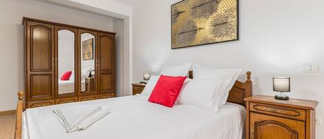A neatly made bed with white bedding and a red pillow, a wooden wardrobe, a bedside table with a lamp, and a wall-mounted abstract art piece.