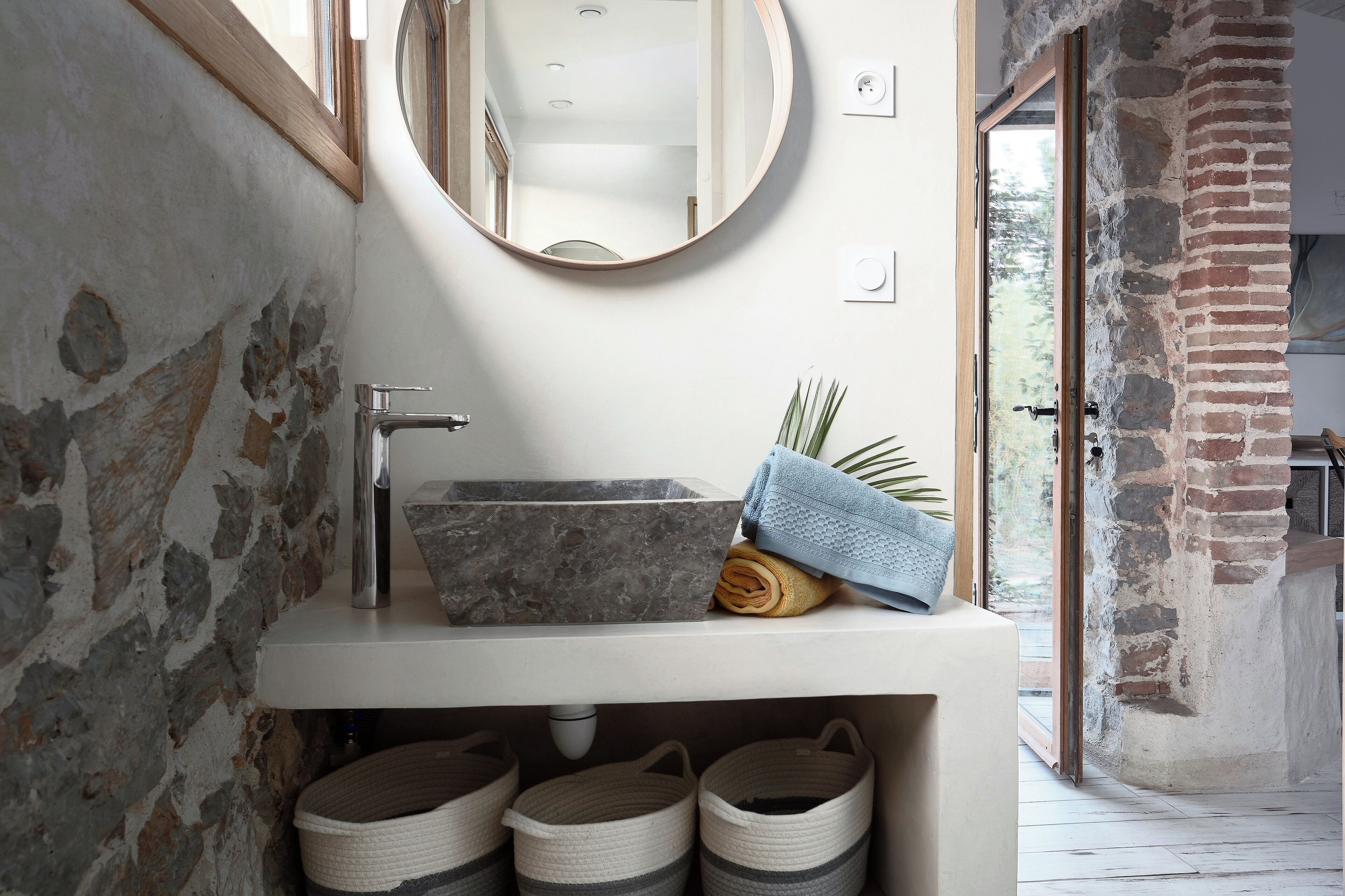 A bathroom with a stone sink, a round mirror, and a view of a brick wall.
