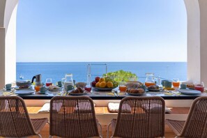 A breakfast table with a view of the sea, wicker chairs, and a variety of breakfast items.