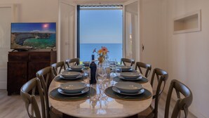 A dining area with a round table set for six, a television on the wall, and a view of the sea through an open door. - Villa Horama - Waterfront with Infinity Pool (Siracusa)