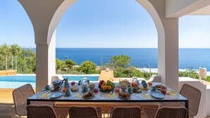 A dining table set for breakfast with a view of the sea, surrounded by wicker chairs, overlooking a swimming pool. - Villa Horama - Waterfront with Infinity Pool (Siracusa)