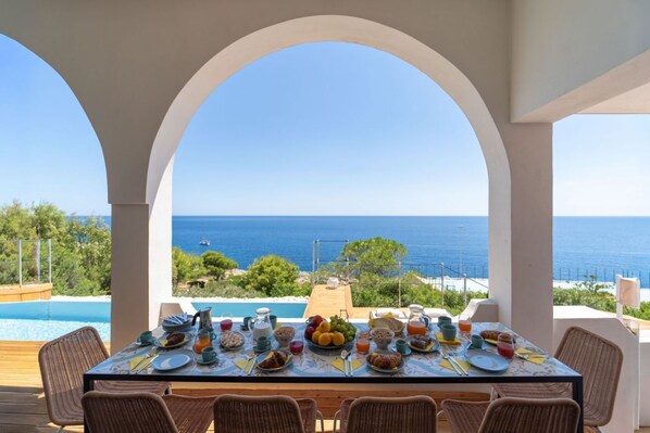 A dining table set for breakfast with a view of the sea, surrounded by wicker chairs, overlooking a swimming pool.