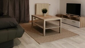 A living room with a wooden ceiling, a sofa, a coffee table with a plant, a TV on a wooden stand, and a window with curtains. - MAISONETTE PROVENCALE (Le Thor)