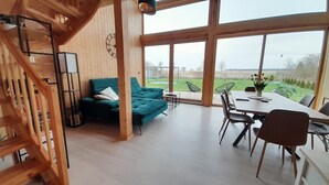 A spacious room with a large window, a green sofa, a dining table with chairs, and a staircase. - Villa Leone - spacious, breathtaking in harmony with nature (Nowe Warpno)