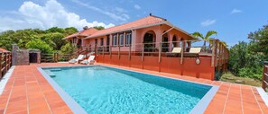 A poolside area with a swimming pool, lounge chairs, and a building with a red-tiled roof.