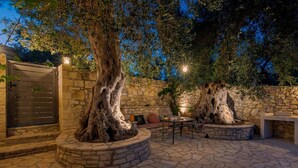 Un espace extérieur aménagé en terrasse, avec un sol en pierre, deux imposants troncs d’arbres, un mur en pierre et un portail en métal.