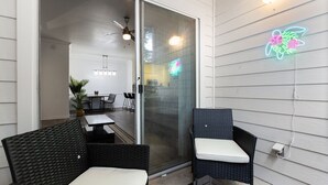 Una terrazza esterna con mobili in vimini, una porta scorrevole in vetro che dà su una cucina e un neon con un fiore sulla parete.