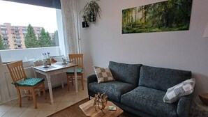 A living room with a sofa, a coffee table, a dining table with chairs, a window with a view of buildings, and a painting on the wall. - "Waldgeflüster" apartment with Harz romance. (Altenau)