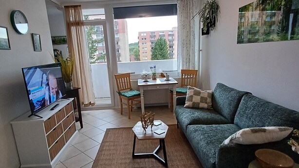 A modern living room with a sofa, coffee table, and TV. There is a dining area with a table and chairs, and a view of buildings outside. - "Waldgeflüster" apartment with Harz romance. (Altenau)