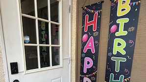 Una puerta blanca con un ventanal, un felpudo con la palabra 'home' y dos pancartas verticales que dicen 'HAPPY BIRTHDAY'.