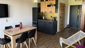 A compact kitchen with wooden cabinets, a black refrigerator, and a small island with a sink. There is a dining table with chairs and a hammock chair.