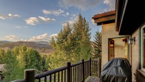 Una terraza de madera con una parrilla de barbacoa, con vistas a un paisaje montañoso con árboles y cielo despejado.