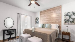 Quarto com cama, dois banquinhos, escrivaninha, cadeira, mesa de cabeceira, ventilador de teto e janela com cortinas.
