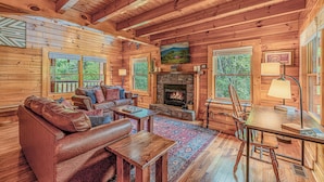 A cozy log cabin interior with a fireplace, leather sofa, wooden dining table, and large windows. - Romantic Cabin • Hot Tub • Pond View • Easy Roads • 5 Min to Parkway (Pigeon Forge)
