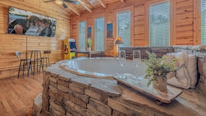A wooden cabin interior with a stone fireplace, a hot tub, and a bar area. - Romantic Cabin • Hot Tub • Pond View • Easy Roads • 5 Min to Parkway (Pigeon Forge)
