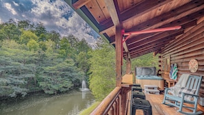 A wooden cabin with a deck overlooking a forested area and a river. - Romantic Cabin • Hot Tub • Pond View • Easy Roads • 5 Min to Parkway (Pigeon Forge)