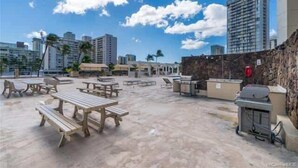 Un espace extérieur aménagé en terrasse, avec des tables de pique-nique et un barbecue, entouré d’immeubles de grande hauteur et de palmiers.