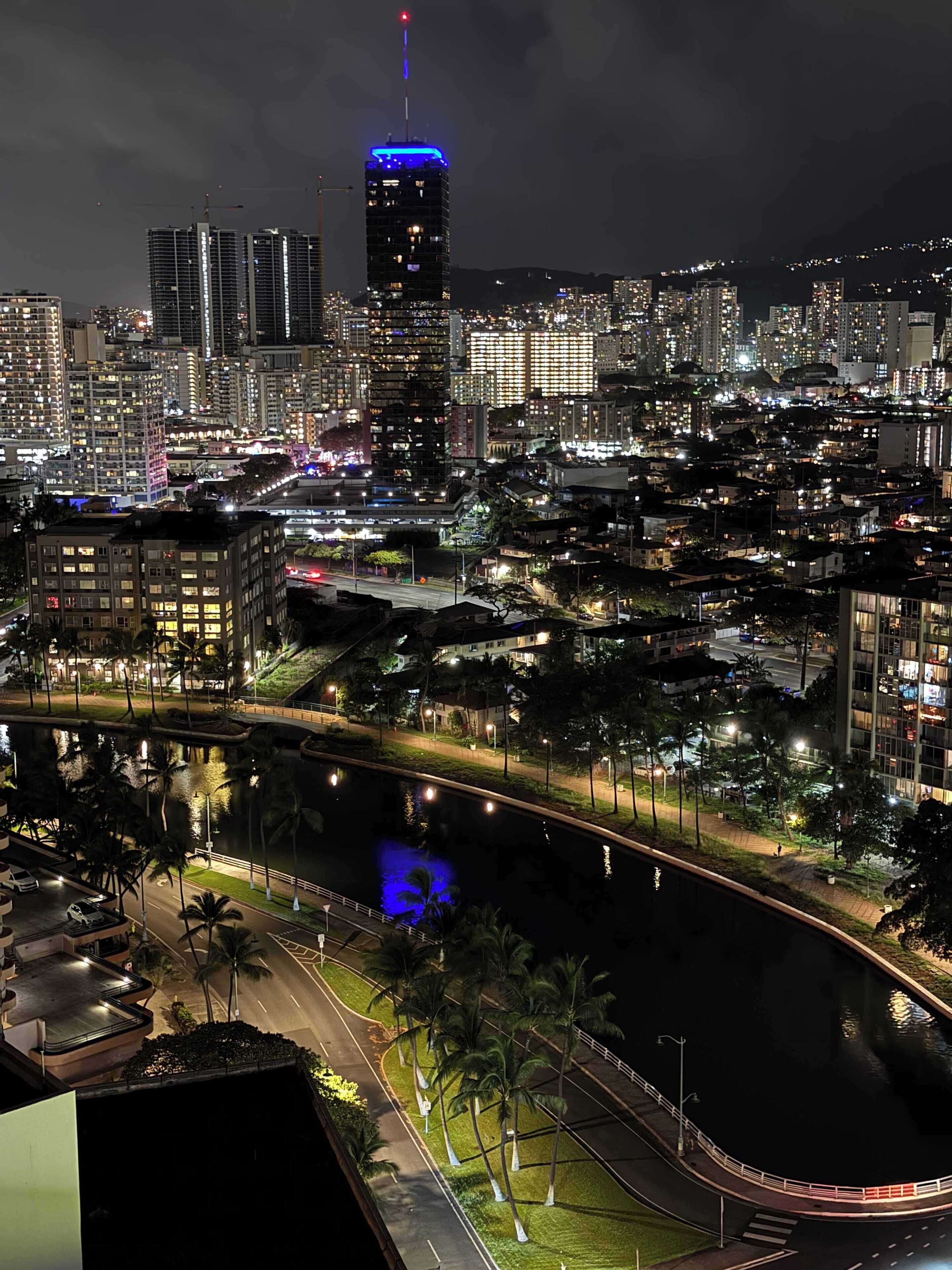 A nighttime cityscape with a prominent building featuring a blue-lit top, a river running through the city, and a bridge connecting two areas.