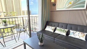 A modern living room with a dark sofa, a small table, and a potted plant. There is a balcony with a view of the city and tall buildings. - Waikiki Ocean View Studio, 3 Beds, Block to Beach (Honolulu)