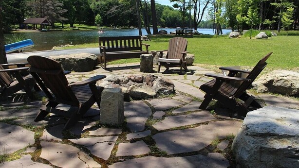 A stone patio with Adirondack chairs and a wooden table, overlooking a lake.