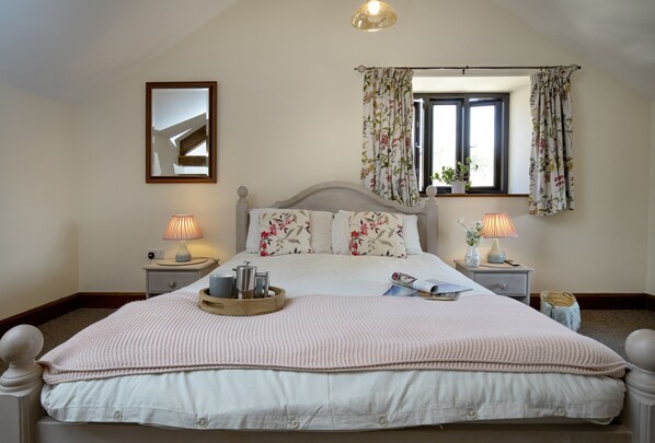 Een slaapkamer met een groot bed, twee nachtkastjes, een raam met bloemen gordijnen, een spiegel aan de muur en een plafondlamp.