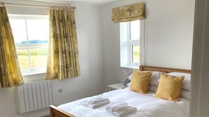 Une chambre avec un lit en bois, une literie blanche et deux oreillers jaunes. On y trouve une fenêtre avec des rideaux, un radiateur et une petite table de chevet avec une lampe.