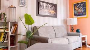 A living room with a grey sofa, a side table with a lamp, a bookshelf, and a large green plant. - LtPanella Jacqueline (Roma)