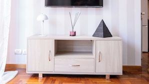 A room with a wooden herringbone floor, a white striped wall, a mounted flat-screen TV, a white desk with a lamp, and a small decorative item. - LtPanella Jacqueline (Roma)