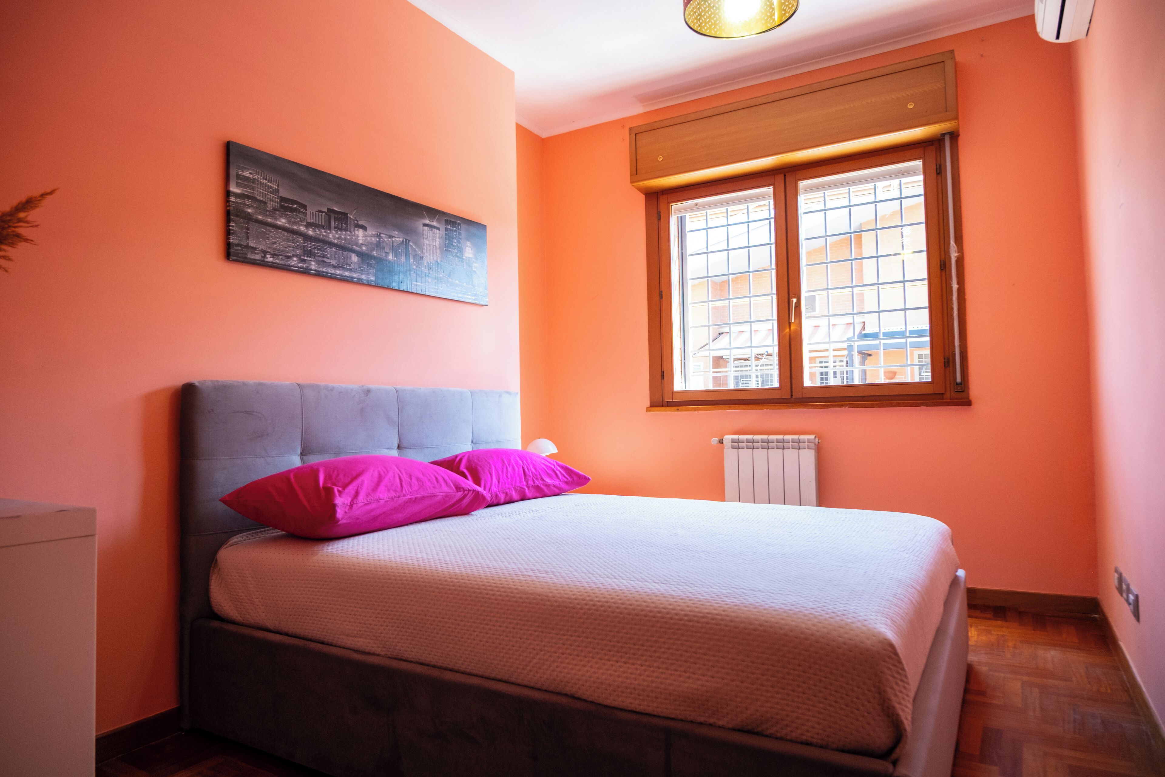 A bedroom with a bed, a radiator, a window, and a cityscape painting.