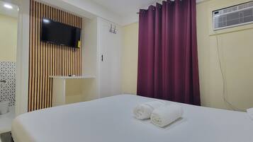 Habitación de hotel con cama, televisor montado en la pared, armario y ventana con cortinas.