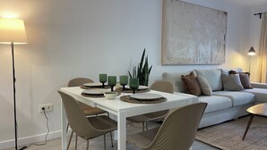 Een eethoek met een witte tafel, geweven stoelen en groene kandelaars. Een bank en een schilderij staan op de achtergrond.
