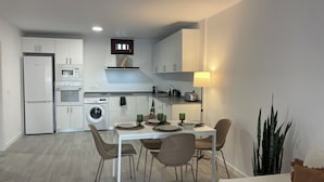 Een compacte keuken met een koelkast, magnetron, oven en wasmachine. Er is een eettafel met stoelen en een lamp.