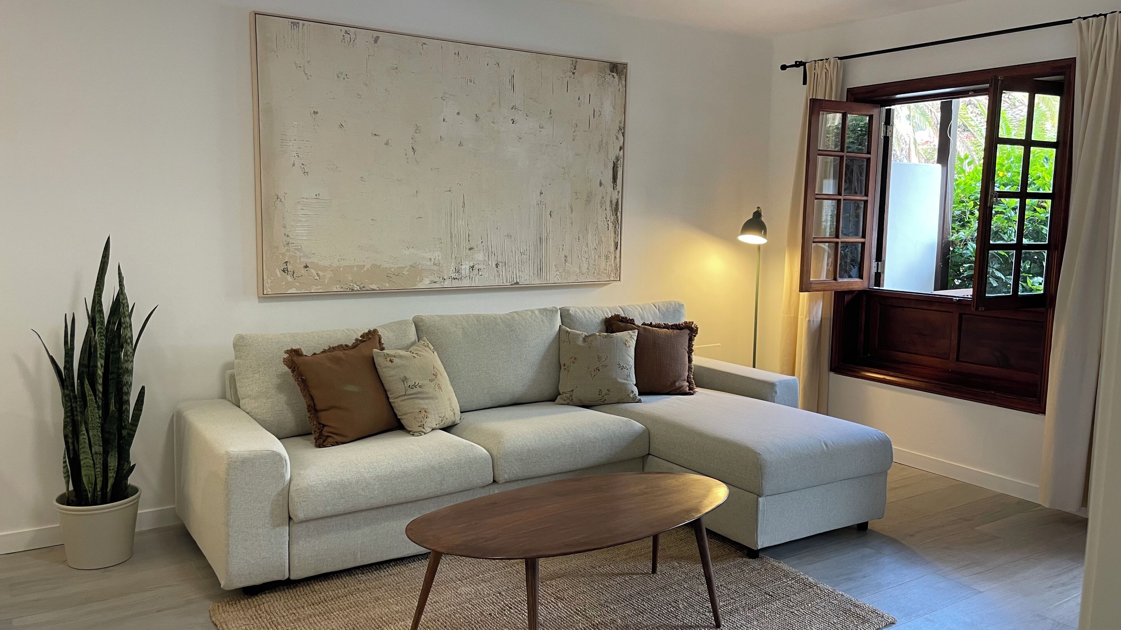 Een woonkamer met een lichtkleurige bank, een salontafel, een groot abstract schilderij, een potplant en een raam met gordijnen.