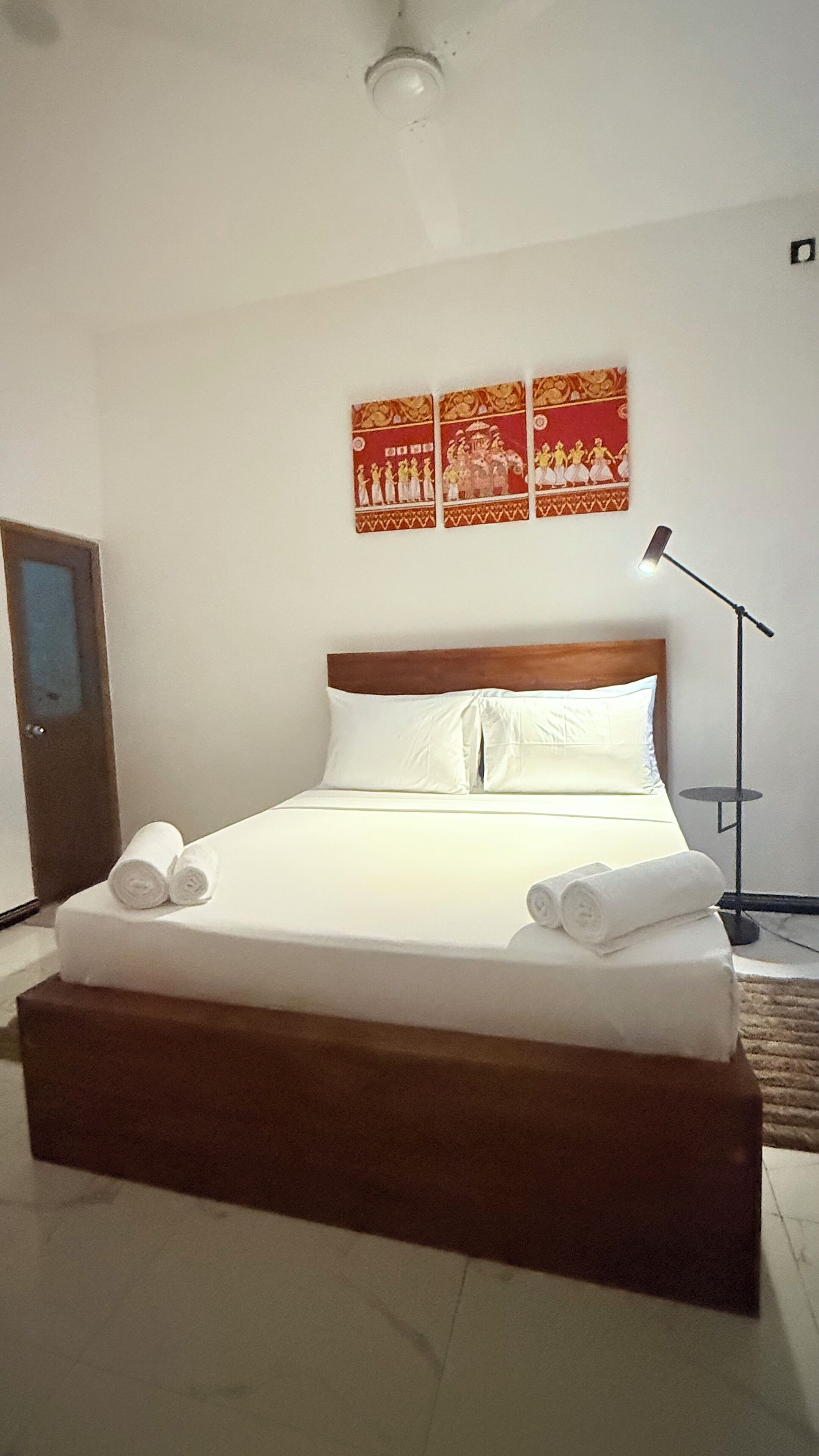 Una cama bien hecha con sábanas blancas y toallas enrolladas, cabecera de madera y un cuadro en la pared que representa figuras tradicionales.