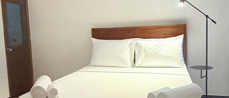 Una cama bien hecha con sábanas blancas y toallas enrolladas, cabecera de madera y un cuadro en la pared que representa figuras tradicionales.