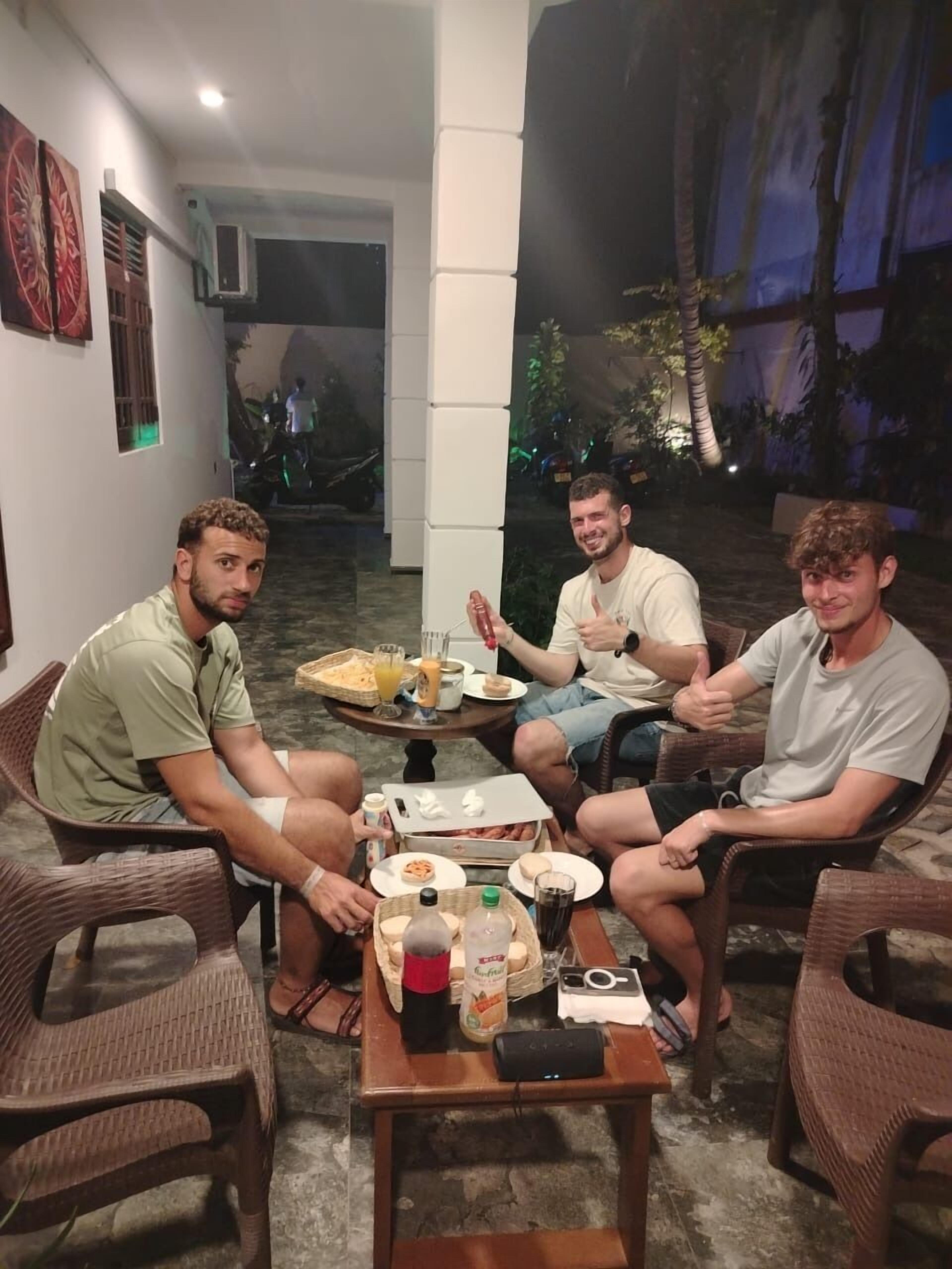 En grupp människor sitter runt ett bord med mat och dryck utomhus, med utsikt över träd och en byggnad i bakgrunden.
