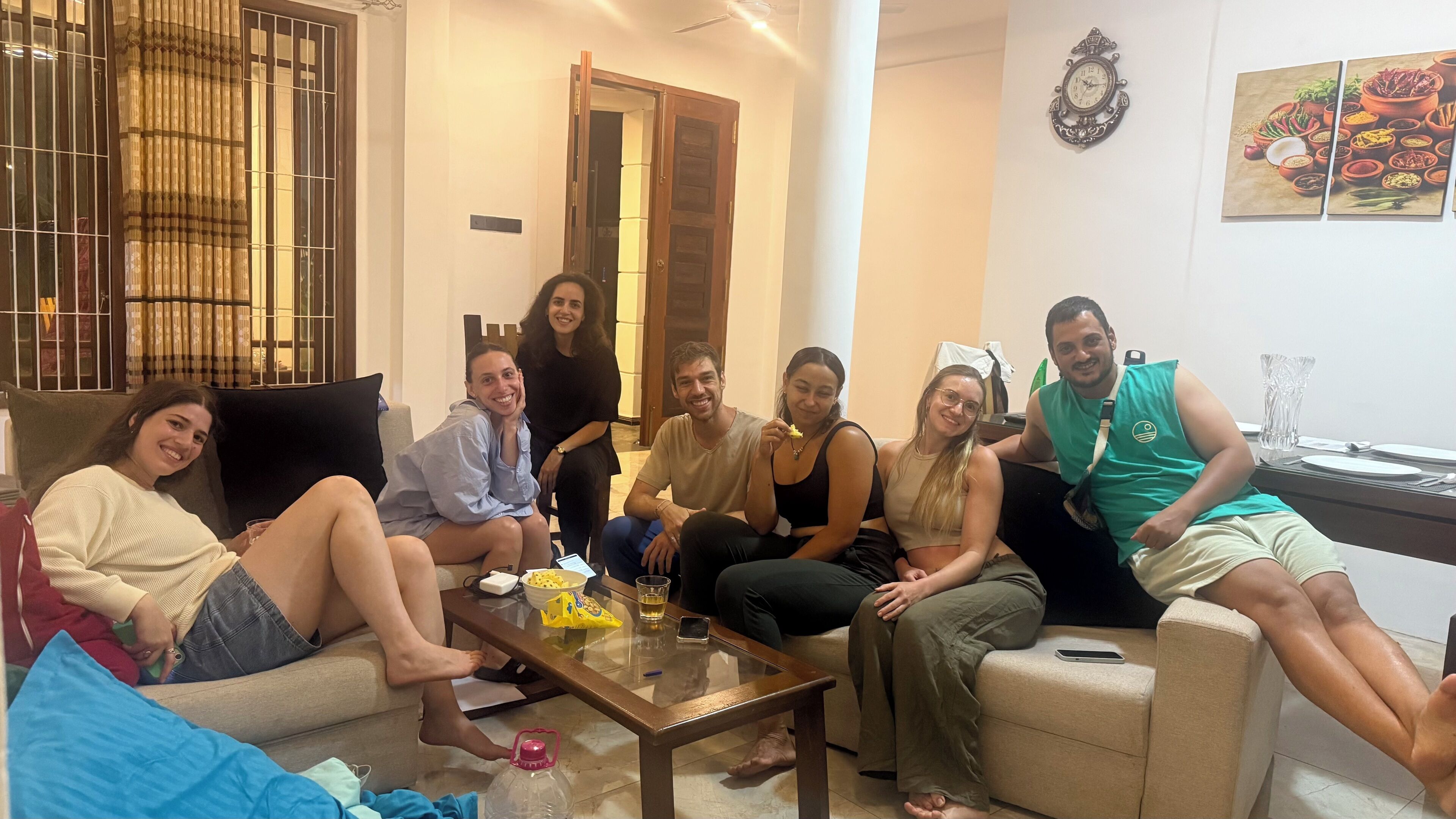 A group of people sitting on a couch and chairs in a well-lit room with a glass coffee table, a door, and a wall clock.