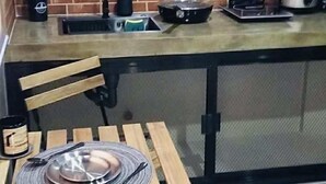 Pieni keittiö, jossa on tiiliseinä, pesuallas, liesi ja pöytä sekä yksi tuoli.