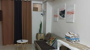 A small, well-lit living area with a sofa, a small round table, and a white bench. There is a window with curtains, a plant, and artwork on the wall. - Cozy City Stay in Baguio (Baguio City)