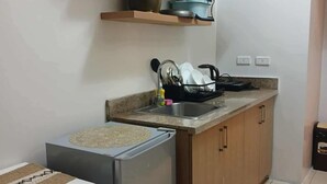 A compact kitchen with a refrigerator, a sink, and wooden cabinets. There are shelves above the counter holding kitchenware and a potted plant. - Cozy City Stay in Baguio (Baguio City)