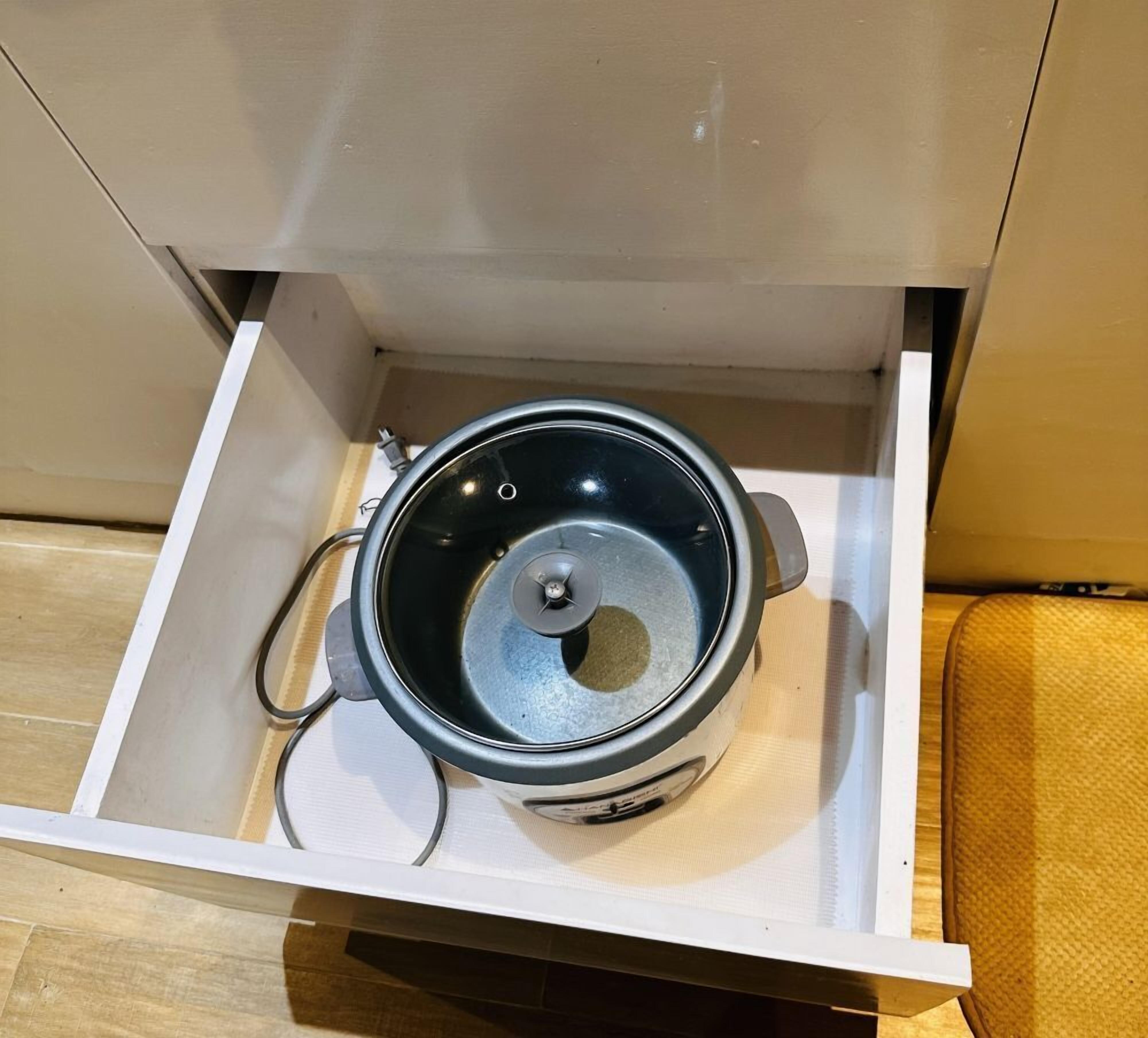 A kitchen drawer containing a stainless steel pot with a lid, a power cord, and a control panel.
