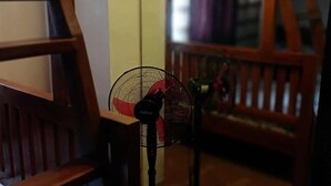 Une pièce meublée de meubles en bois, notamment une chaise et un lit. On aperçoit un ventilateur sur pied au premier plan.
